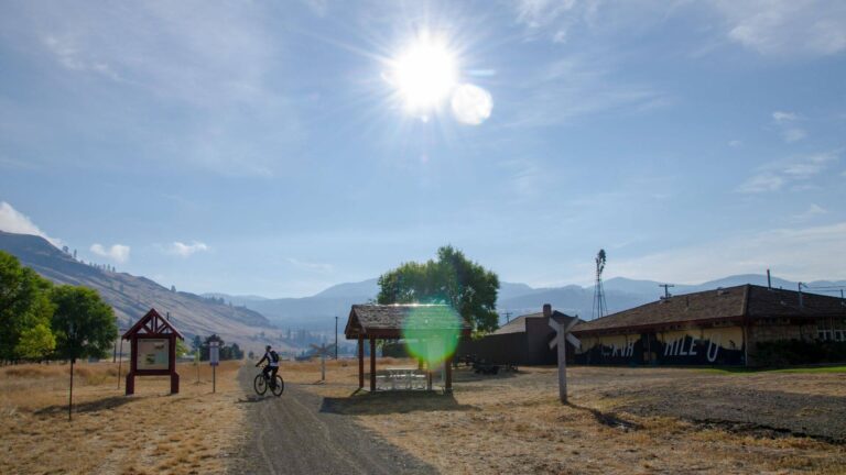 A person rides a bicycle on a gravel path through a sunny, rural landscape with mountains in the background, a picnic shelter, and trees nearby under a bright blue sky.