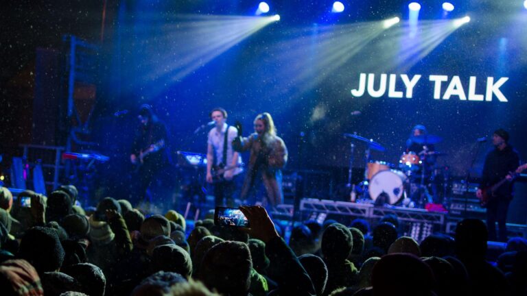 A band performs on stage under blue lights with the name JULY TALK displayed behind them. The audience watches and some people hold up phones to record the concert.