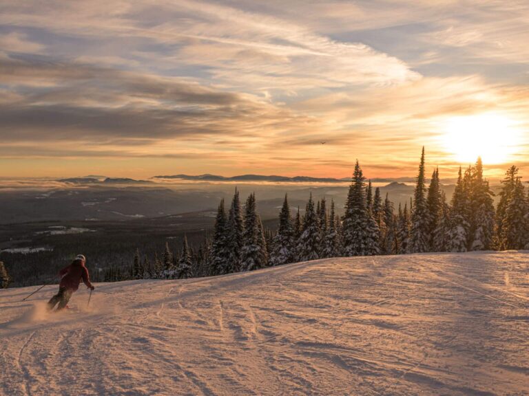 A skier glides down a snowy slope at sunset, surrounded by snow-covered pine trees and distant mountains under a partly cloudy sky—a perfect scene for those seeking inspiration on where to go for their next adventure.