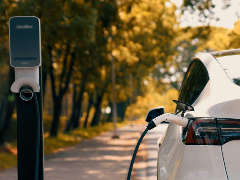 A white electric car is parked on a tree-lined street, plugged into a charging station, as sunlight filters through the leaves—an inspiring scene that echoes the positivity of 7 Affirmations.