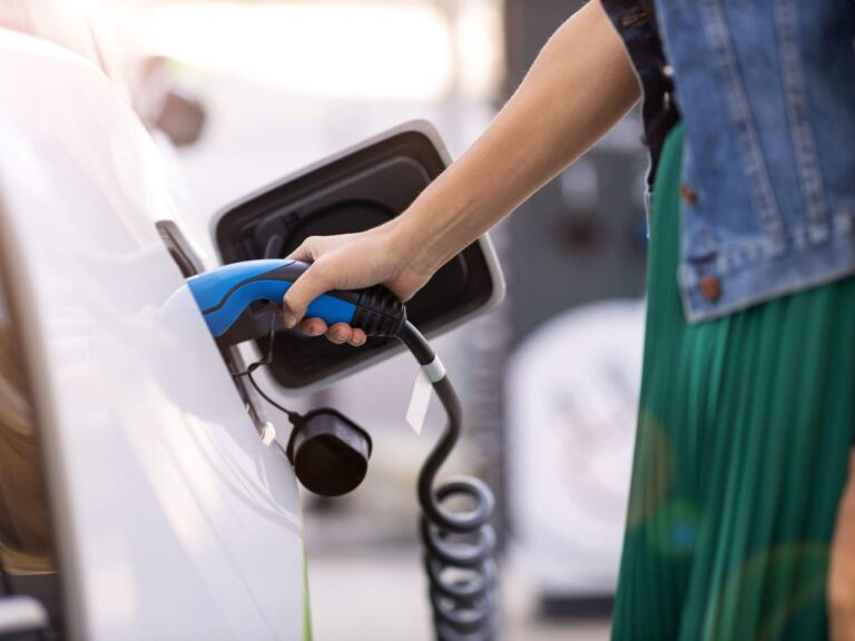 A person holds a blue electric vehicle charging plug, connecting it to a white car’s charging port—powering up with purpose, much like 7 affirmations for positive energy. The person wears a green skirt and denim jacket; the background is out of focus.