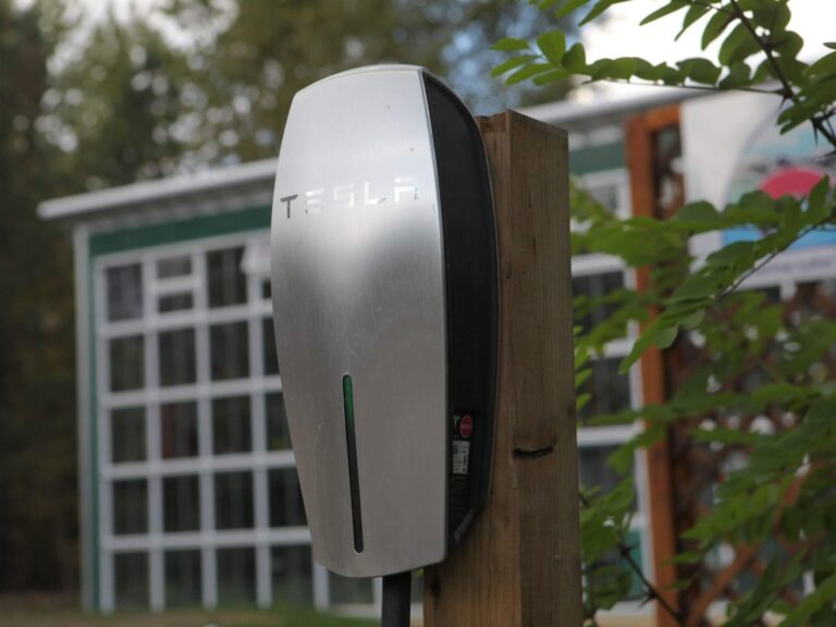 A Tesla electric vehicle wall charger is mounted on a wooden post outdoors, with trees and a building in the blurred background—an inspiring setup for those who believe in progress and the power of 7 affirmations to shape the future.