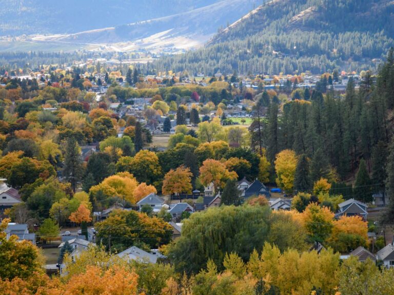 A scenic view of a town surrounded by trees with autumn foliage in shades of orange, yellow, and green, set against forested hills and distant mountains under a partly cloudy sky.
