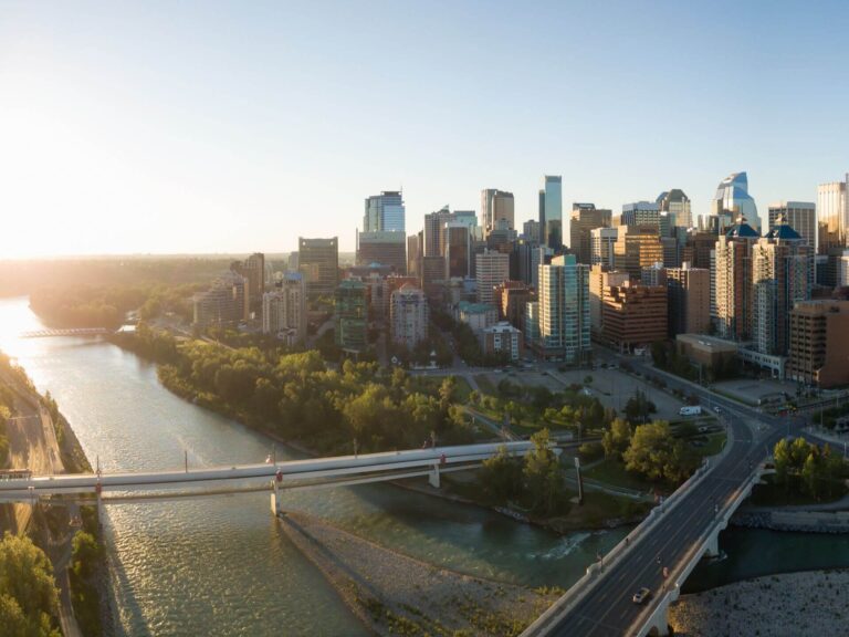 Aerial view of a river flowing beside a modern city skyline with tall buildings, green trees, and several bridges crossing the water under a clear sky at sunset, inspiring calm like daily 7 Affirmations.