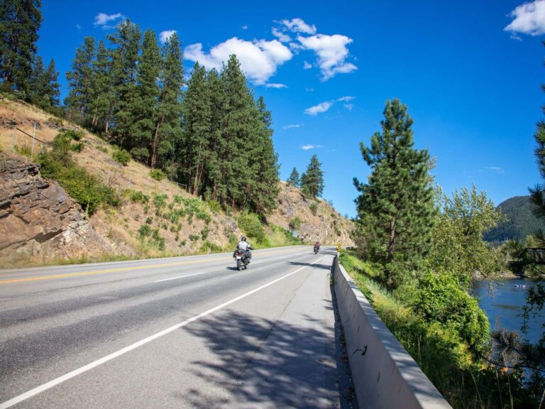 Two motorcyclists ride along a sunny, winding highway bordered by trees and rocky hills, embracing the journey with 7 affirmations in mind, as a clear blue sky and forested riverbank stretch out to the right.