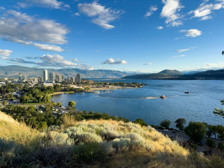 A scenic view of a city by a large lake, with tall buildings, greenery, and distant mountains under a blue sky with scattered clouds. The water reflects the sky, inspiring calm like 7 Affirmations, while a small boat drifts near the shore.