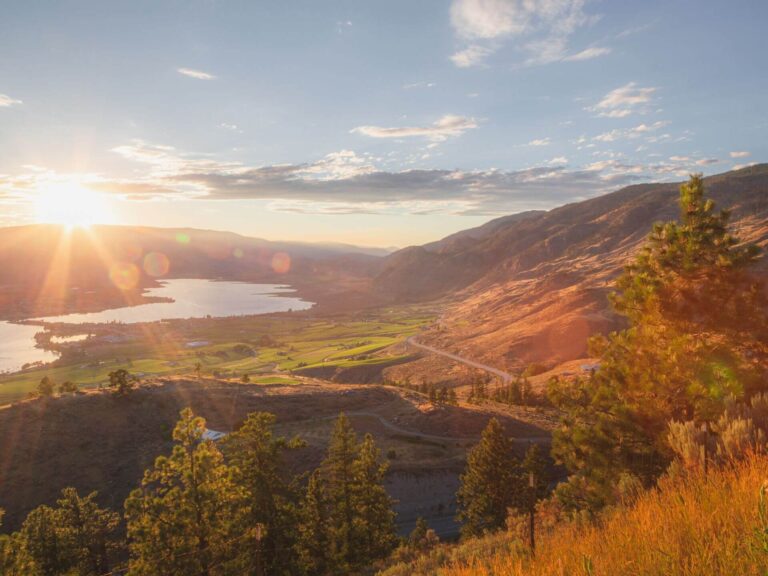 A scenic view of a sunlit valley with a lake, green fields, and winding roads offers tranquility—perfect for reflecting on 7 Affirmations—surrounded by forested hills and mountains under a partly cloudy sky at sunrise or sunset.