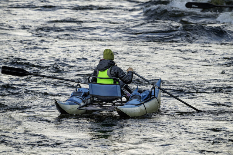 Person in a green hat and life vest rows a small inflatable cataraft on a flowing river, surrounded by water with visible ripples and waves.