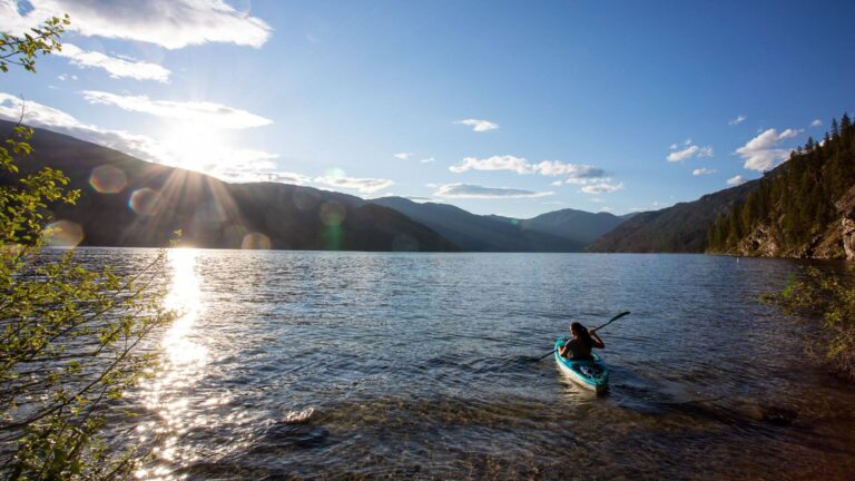 A person paddles a kayak on a clear lake surrounded by mountains and forest, with the sun shining low in the sky and reflecting on the water.