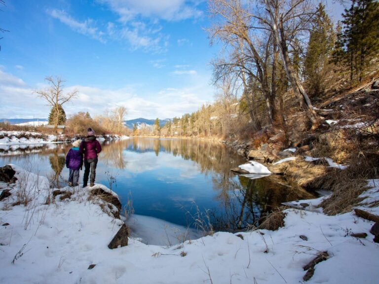 Two people in winter jackets stand by a calm, snow-covered, pet friendly riverbank, surrounded by leafless trees and evergreens, with mountains and a clear blue sky in the background.