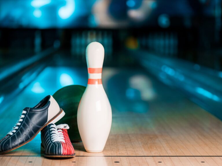 A pair of bowling shoes, a green bowling ball, and a white bowling pin with red stripes rest on a wooden bowling lane, capturing the classic excitement of bowling amidst blurred lanes and pins in the background.