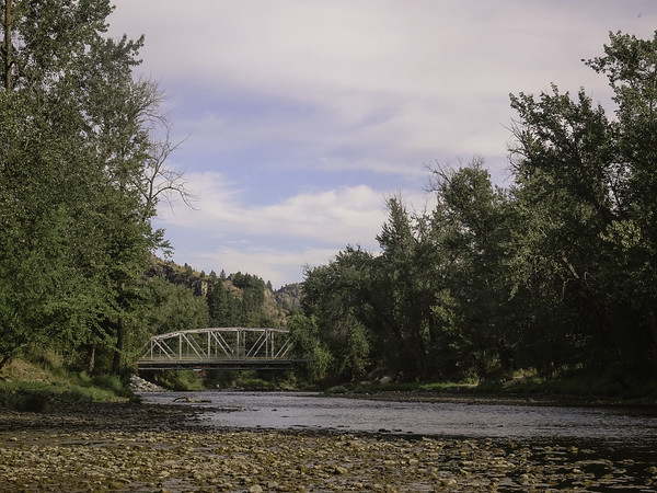 A metal truss bridge spans a shallow, rocky river surrounded by dense green trees under a partly cloudy sky.