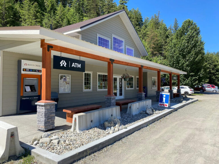 A small building in Boundary Country with an ATM and information booth sign, wooden pillars, benches, and a gravel parking lot, surrounded by green trees on a sunny day. A blue OPEN sign is near the entrance.