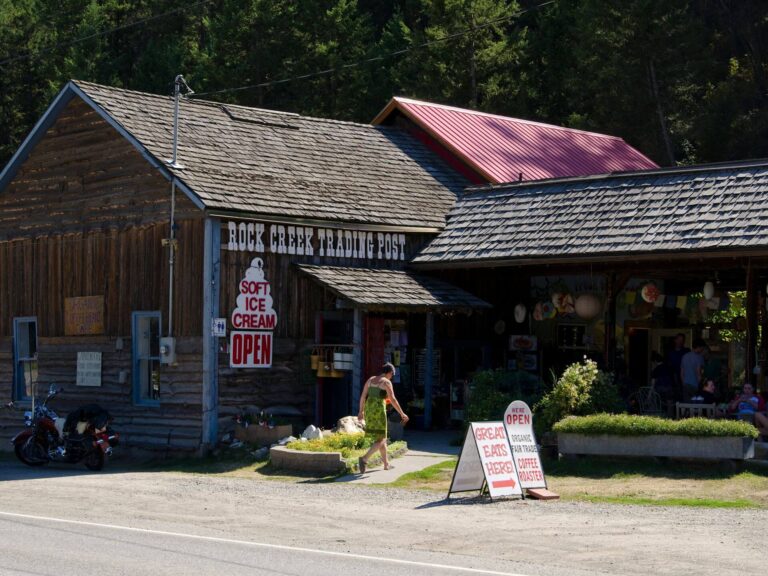 A rustic wooden building with a sign reading Rock Creek Trading Post and Soft Ice Cream OPEN welcomes visitors. Outdoor signs advertise ice cream and food at this pet-friendly spot, with motorcycles parked nearby and trees in the background.
