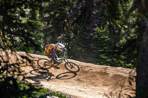 A cyclist wearing a helmet and orange backpack rides a mountain bike on a dirt trail surrounded by green trees and sunlight filtering through the forest.