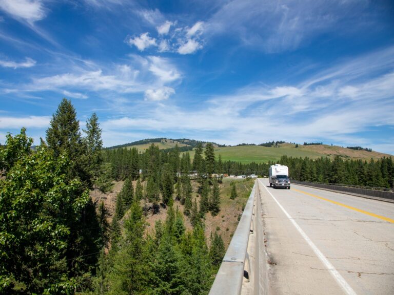 RV crossing a bridge through forested hills under a bright blue sky in Boundary Country, BC