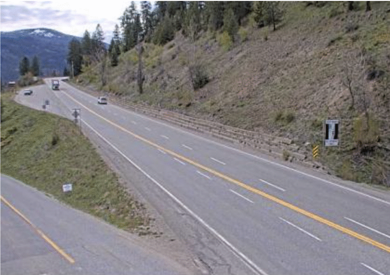 A four-lane highway curves through a hilly, forested area with mountains in the distance. Several cars travel in both directions under a partly cloudy sky, all visible thanks to roadside webcams capturing the scenic drive.