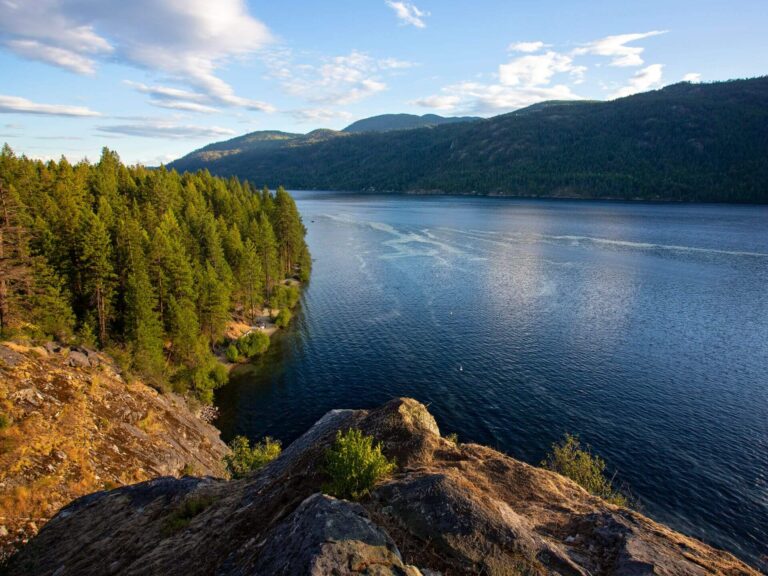 A scenic view of a calm lake bordered by dense pine forests and rocky shorelines, with mountains in the background under a partly cloudy blue sky.