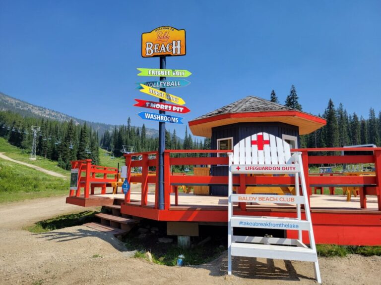 A bright wooden lifeguard hut with a red cross sign stands beside a colorful signpost pointing to Frisbee Golf, Volleyball, Horseshoes, Horseshoe Pit, and Washrooms, surrounded by trees and green hills under a clear blue sky.