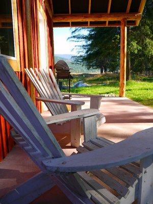 Two wooden Adirondack chairs sit on a sunlit porch with a view of green grass, trees, and distant hills. A barbecue grill is visible near the edge of the porch, next to a rustic wooden building.