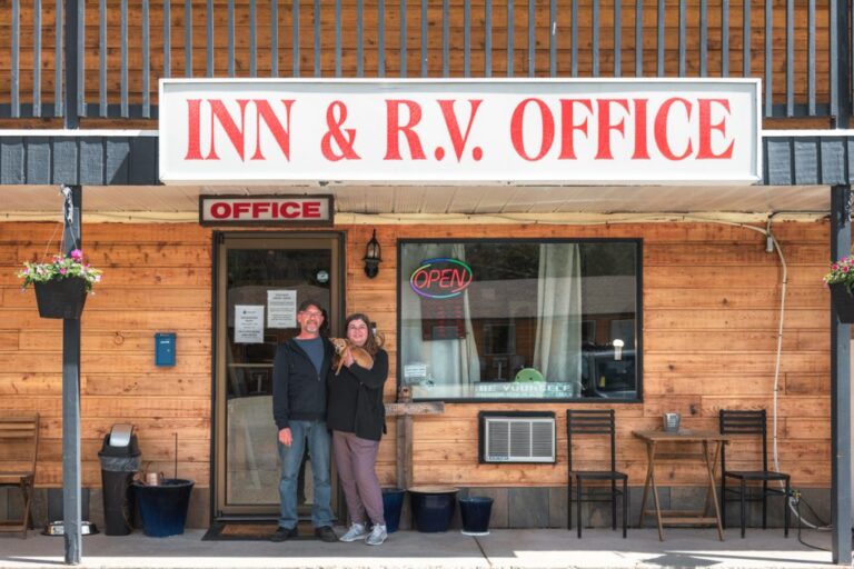 Two people stand smiling in front of a wooden building with a large sign that reads INN & R.V. OFFICE. The office door displays an OPEN sign and there are tables and chairs outside.