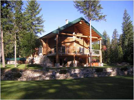 A large log cabin with a spacious covered balcony sits among tall pine trees and a well-manicured lawn, with a stone retaining wall in the foreground.
