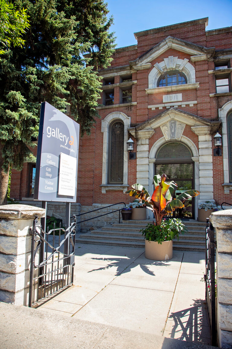 A brick building with arched windows, stone steps, and a potted plant at the entrance. A sign by the gate reads “gallery” and lists art gallery, gift shop, and visitor center. Tall green trees are on the left.