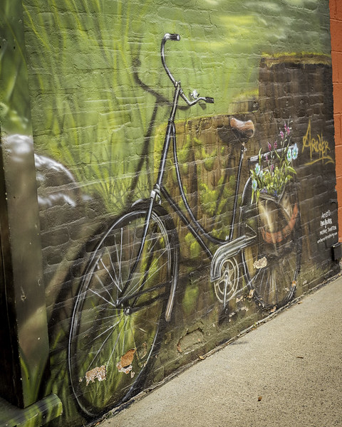 A mural of a vintage bicycle with a basket of flowers painted on a brick wall, blending with a grassy background. The artwork is detailed and realistic, with some areas showing chipped paint.