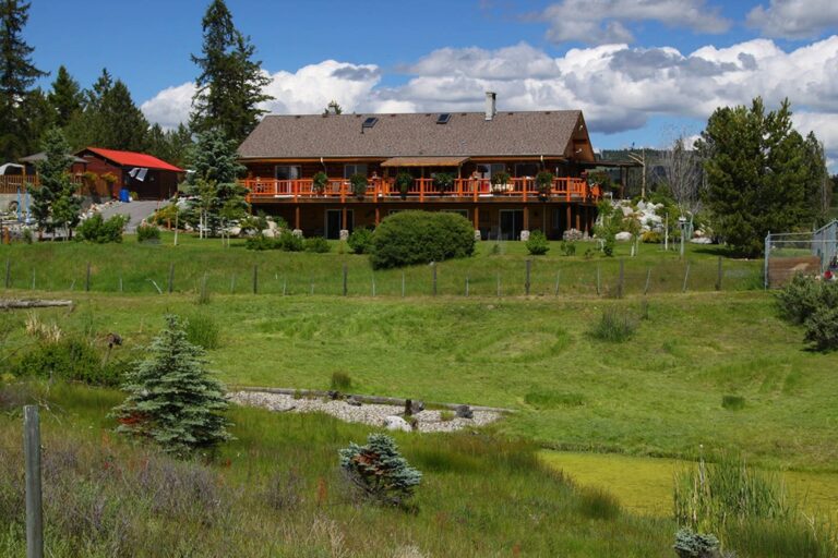 A large wooden house with a wraparound balcony sits on a grassy hill, surrounded by trees, shrubs, and a small pond under a partly cloudy sky.