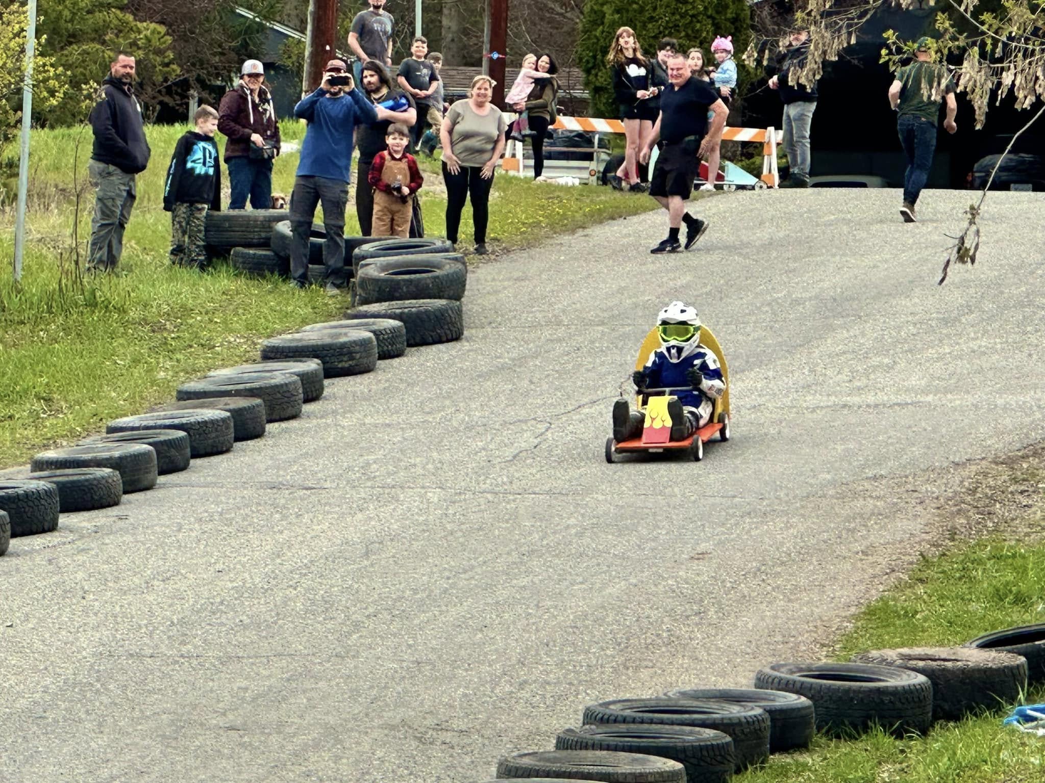 A child wearing a helmet drives a small go-kart down a paved track lined with tires, while a crowd of adults and children watch and take photos from the side.
