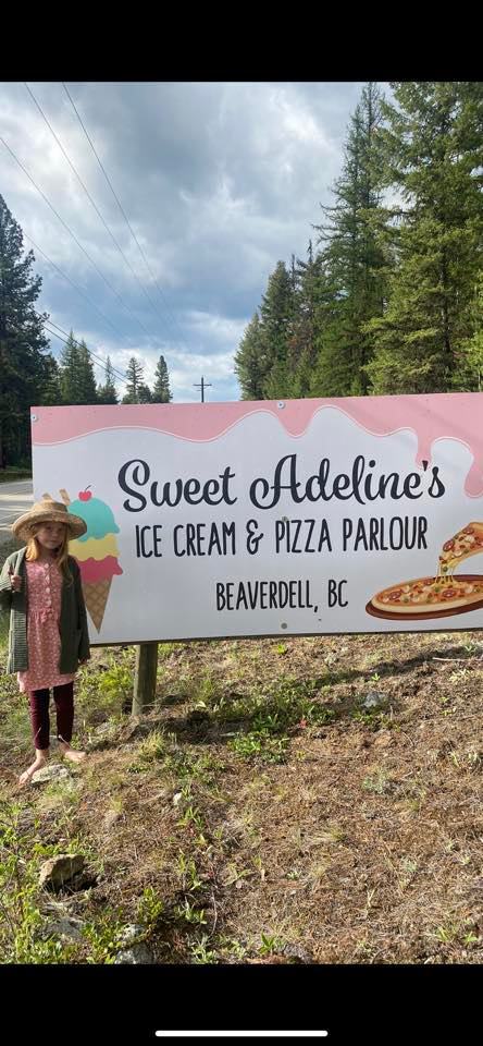 A young girl in a hat and jacket stands beside a large sign that reads Sweet Adelines Ice Cream & Pizza Parlour, Beaverdell, BC, with images of ice cream and pizza, surrounded by trees.
