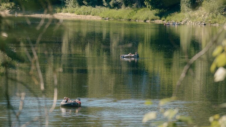 Two people relax in an inflatable raft, and another person floats nearby on a calm, green river surrounded by trees and leafy vegetation, on a sunny day.