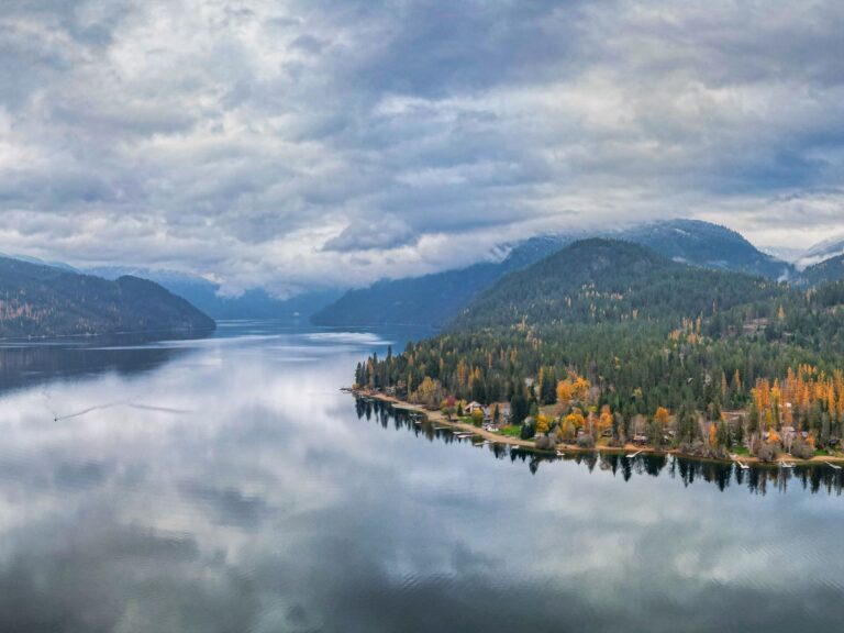 A calm lake in Boundary Country reflects cloudy skies and forested hills, with autumn trees adding orange and yellow hues along the shoreline. Houses nestle among the trees, while mountains fade into the misty distance.