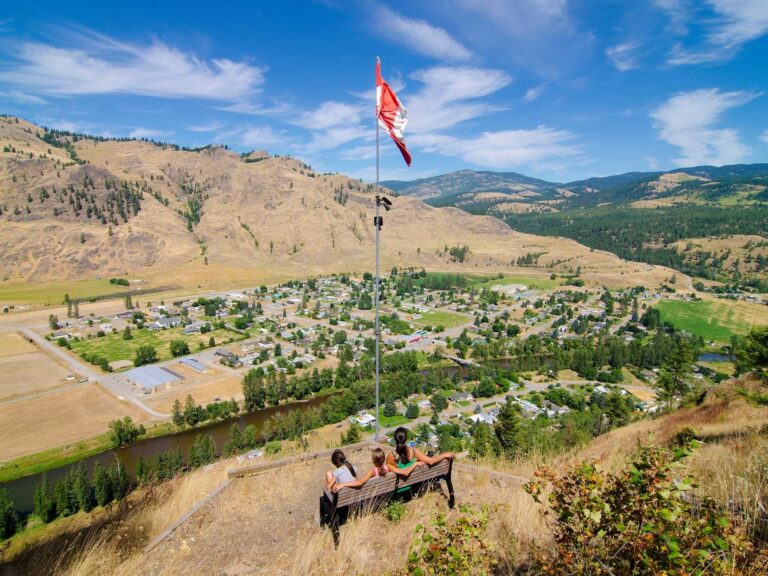 Two people sit on a bench atop a hill in Boundary Country, overlooking a small town with a Canadian flag above them. Rolling hills, scattered houses, and a winding river stretch beneath the bright blue sky and wispy clouds.