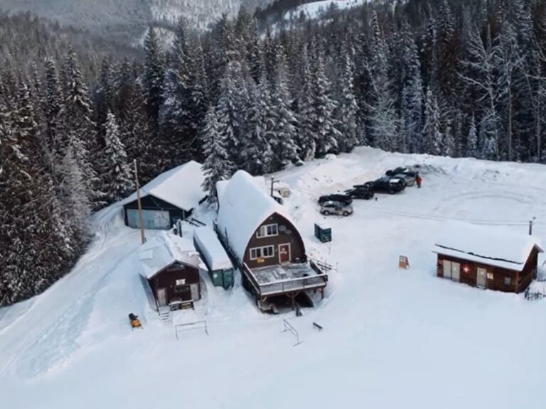 Aerial view of snow-covered cabins and parked cars in Boundary Country, surrounded by dense pine forest. The cabins sit on a cleared area with deep snow and mountains visible in the background.