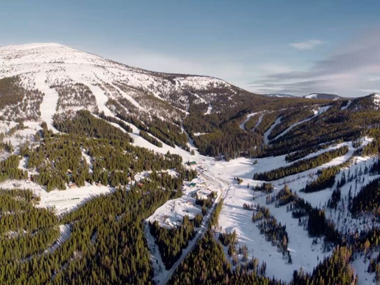 Aerial view of a snow-covered Boundary Country mountain landscape with ski slopes and pine forests. Several trails wind through the trees, and buildings are visible in the valley below under a clear blue sky.
